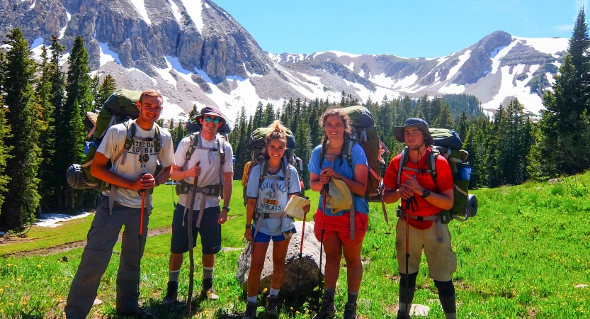 A group of students wearing backpacks pose for a photo in a green meadow. In the background there are snow-capped mountains. 
